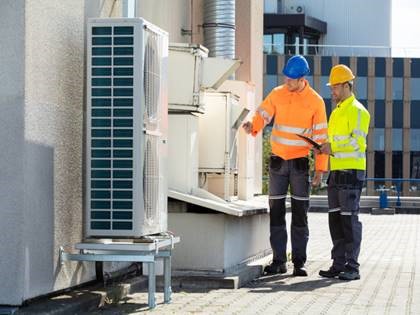 Image of two male construction workers in hard hats and hazard protection clothing inspecting an air conditioning or refrigeration unit